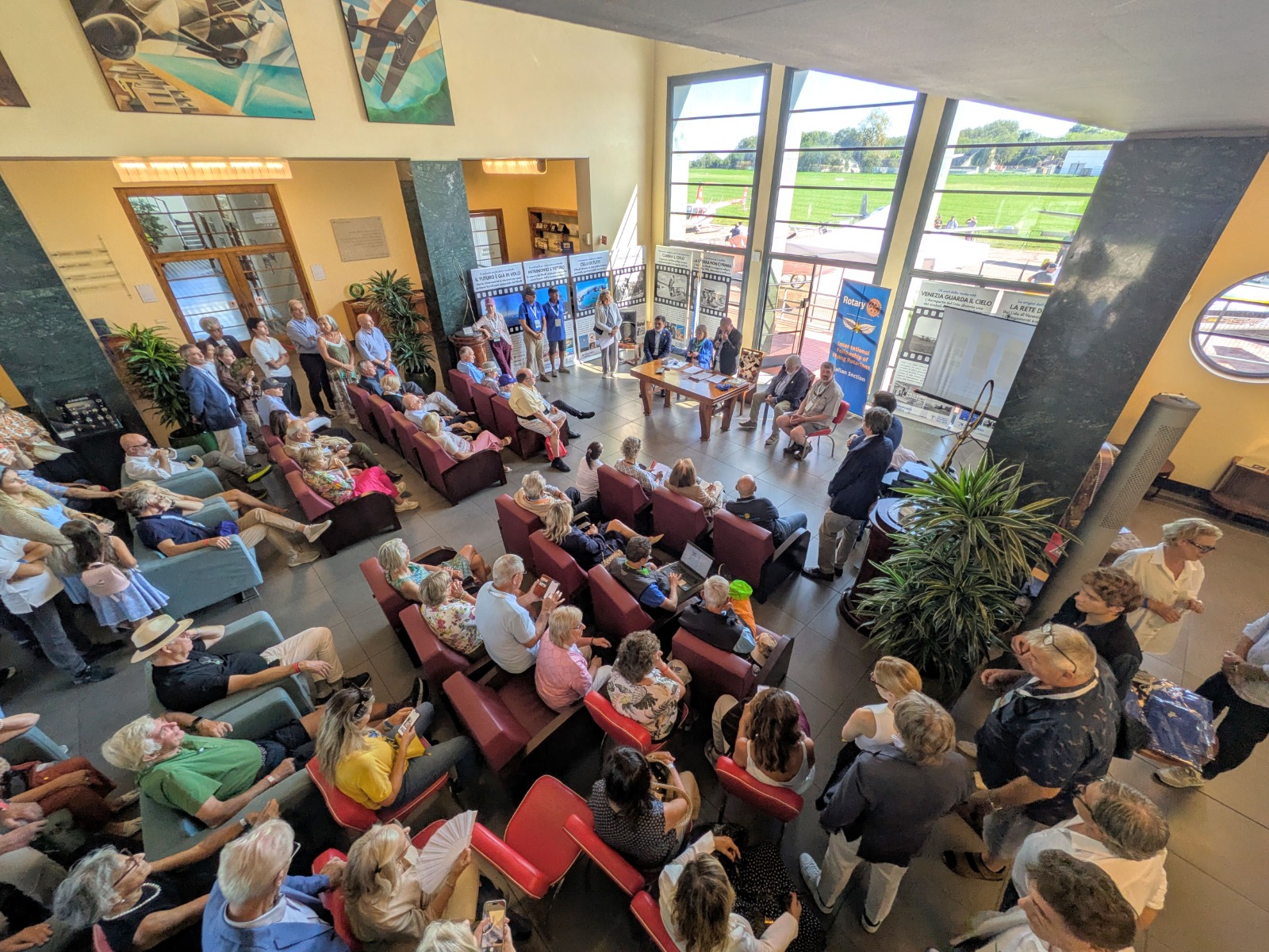 Vista aerea di un evento in una sala con persone sedute e in piedi, partecipanti in attesa di un intervento, tavolo con relatori in primo piano.
