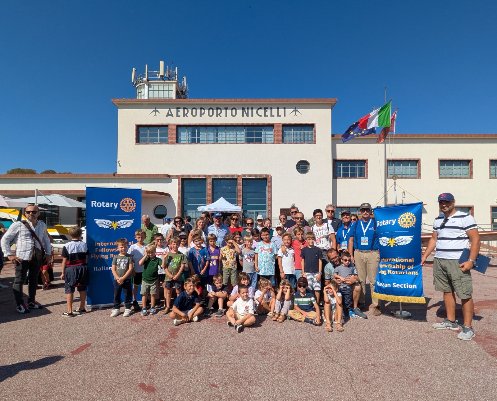 Gruppo di bambini e adulti davanti all'Aeroporto Nicelli, con bandiere e striscioni del Rotary.