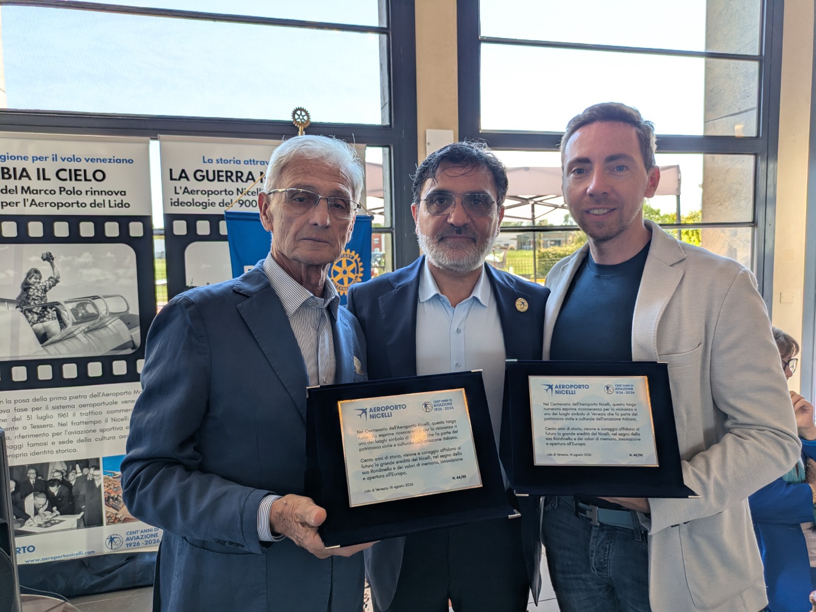 Tre uomini tengono in mano trofei e riconoscimenti durante un evento all'Aeroporto Nicelli, con sfondo informativo.
