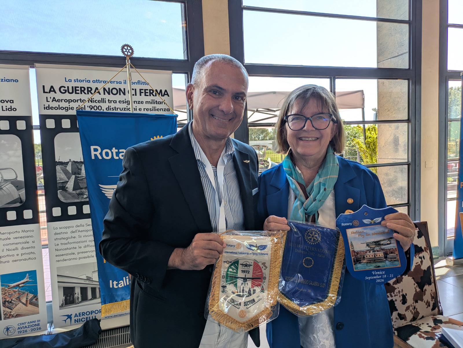 Due persone sorridenti in un evento, entrambe tengono in mano dei gagliardetti del Rotary. Sullo sfondo, banner informativi riguardanti l'aeroporto Nicelli e il Rotary.