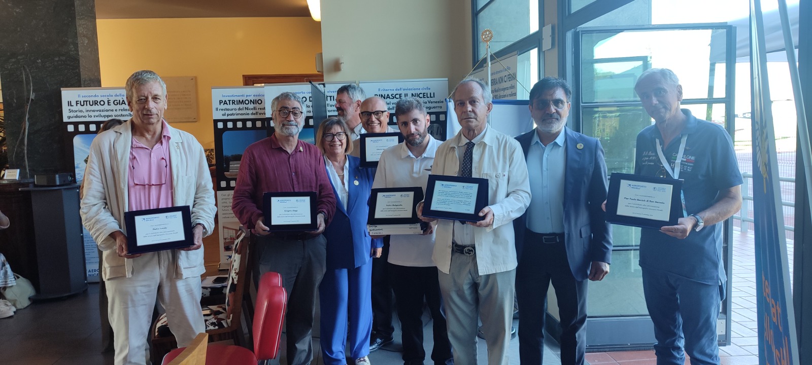 Un gruppo di otto persone ritratto all'interno di un edificio, ciascuna con una targa in mano, durante un evento di premiazione. Sullo sfondo si intravedono dei cartelli informativi.